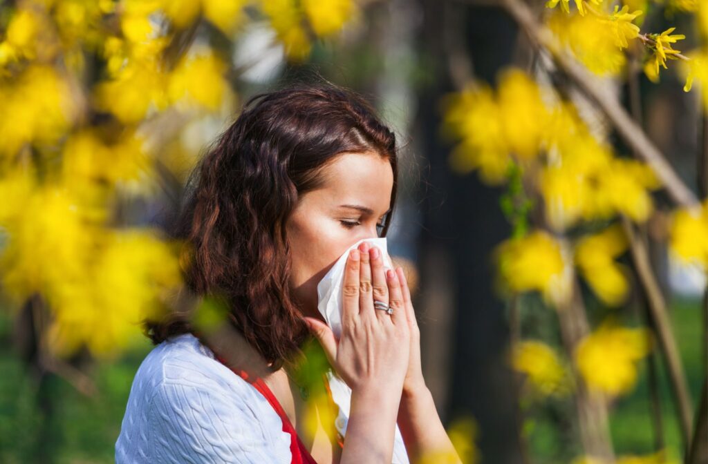 A woman who is standing outside during the spring, blows her nose with a tissue due to allergy symptoms