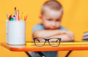 A young boy sitting at a desk and looking at his glasses sitting on his desk in front of him