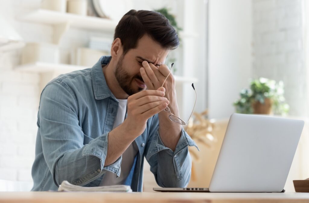 A man working at his computer taking off his glasses to rub his eyes due to meibomian gland dysfunction.