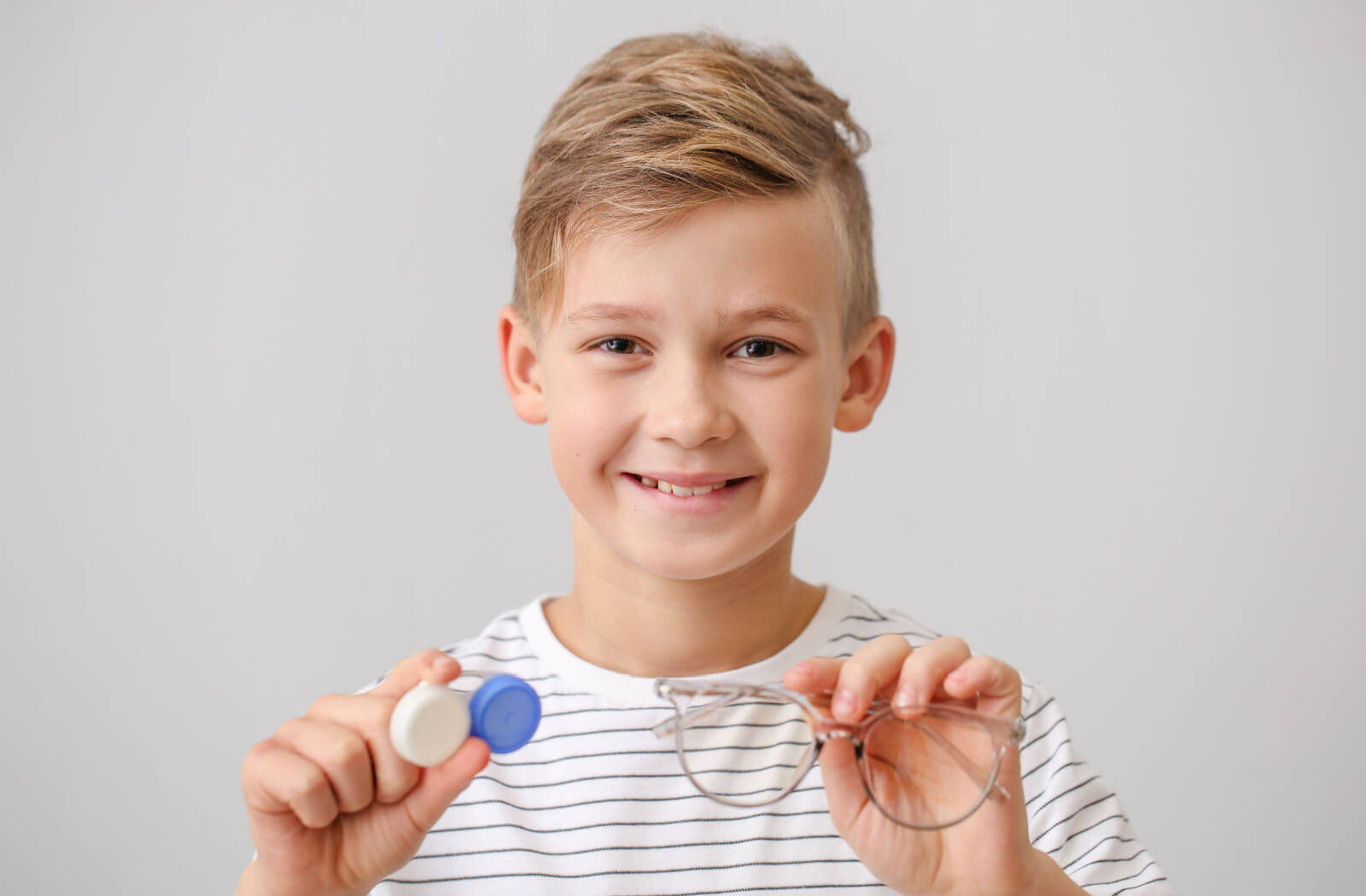 A close-up of a young child smiling while holding up myopia control eyeglasses and contact lenses.