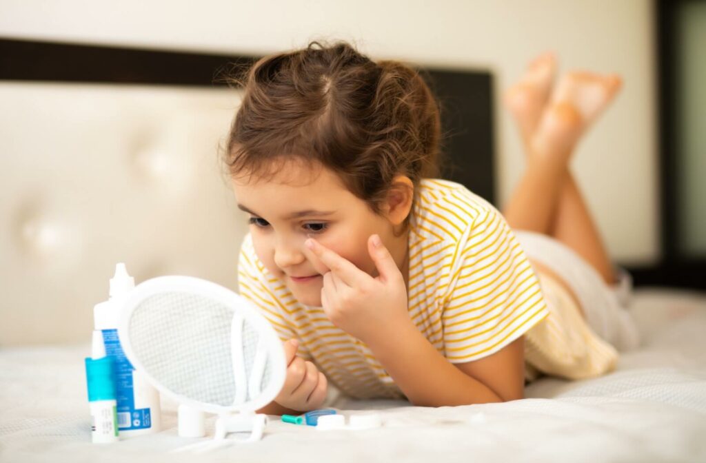Young child applying a contact lens while lying on a bed with lens supplies and a mirror