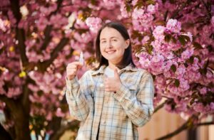 Smiling woman holding eye drops with blooming trees in the background