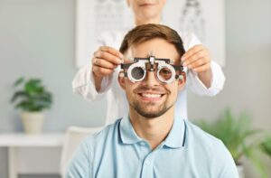 A smiling patient during an eye exam while an optometrist fits their face with a lens phoropter.