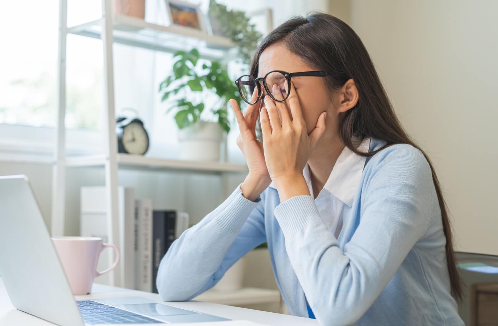 Woman wearing glasses rubbing her eyes while sitting at a desk in front of a laptop, appearing tired or experiencing eye discomfort.
