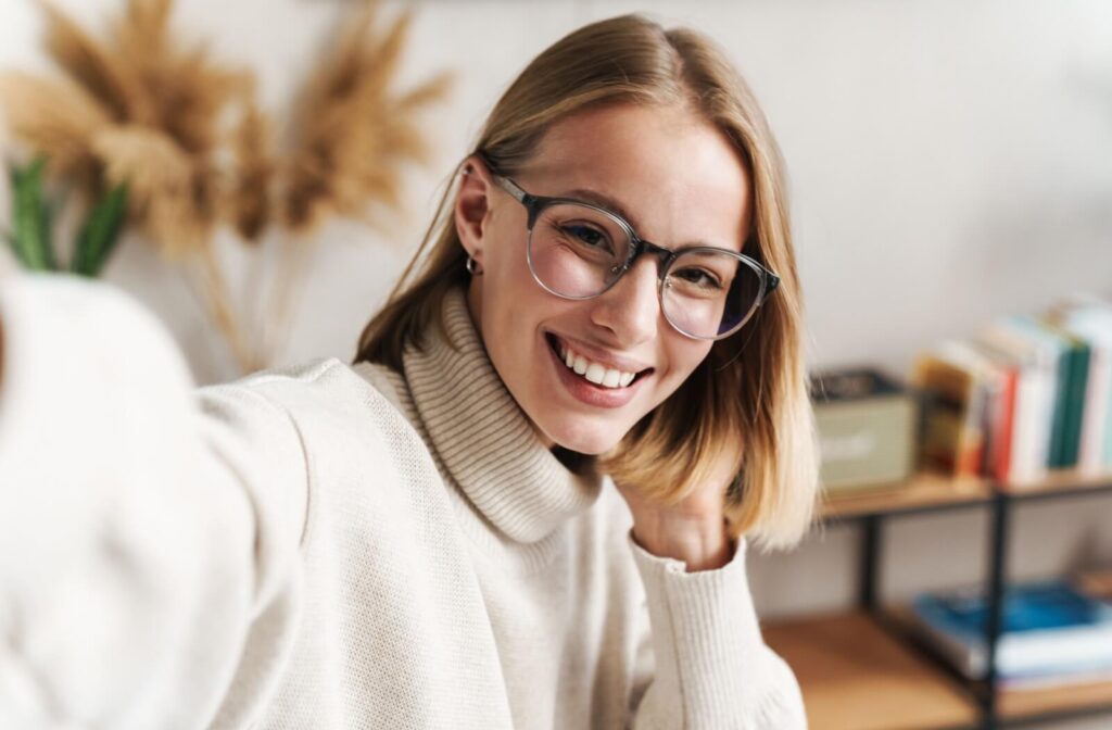 A smiling woman wearing modern, clear-framed eyeglasses takes a casual selfie in a home office