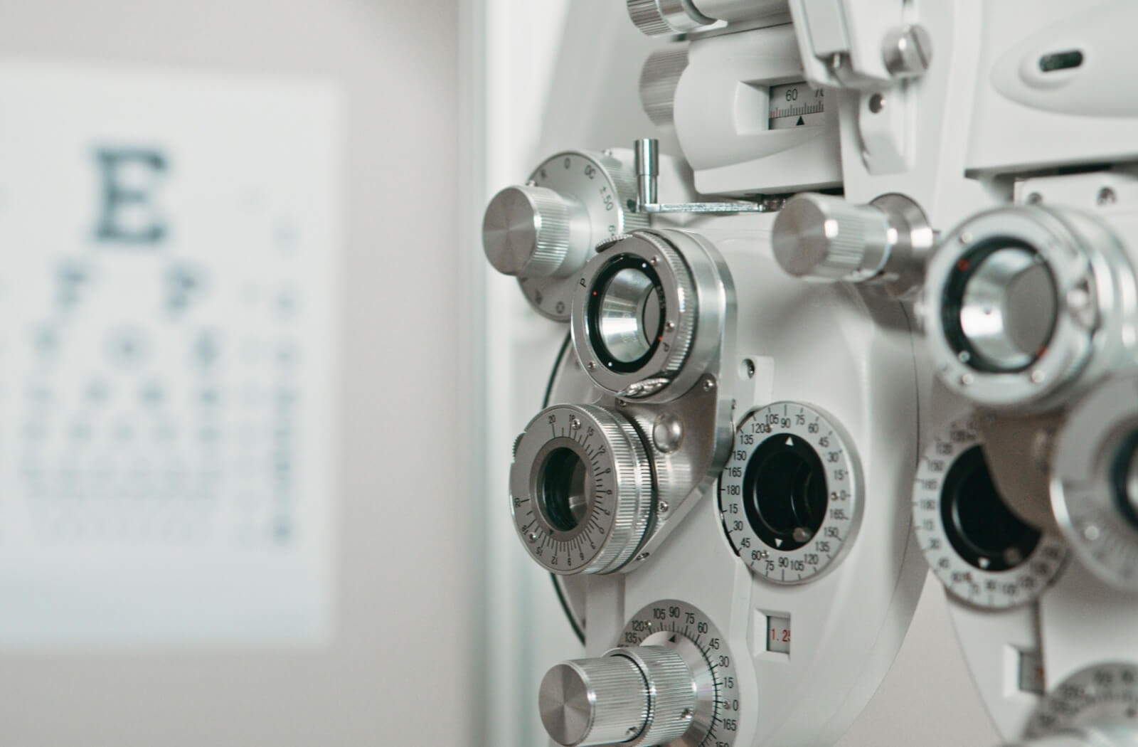 A close-up of a phoropter in an eye exam room with a blurred Snellen eye chart in the background.